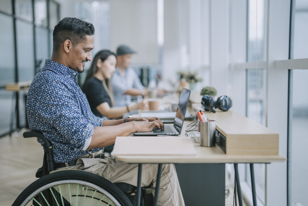 Asian Indian white collar male worker in wheelchair concentrating working in office beside his colleague