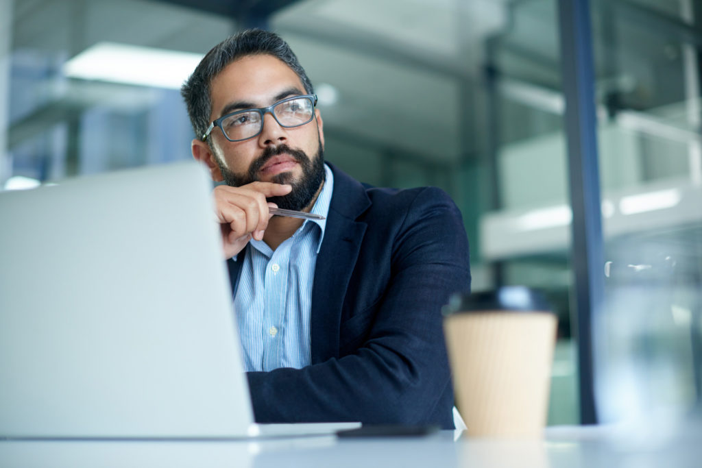 Ein nachdenklicher Geschäftsmann vor dem Laptop, der über Critical Thinking und Reflexion im Arbeitskontext nachdenkt.