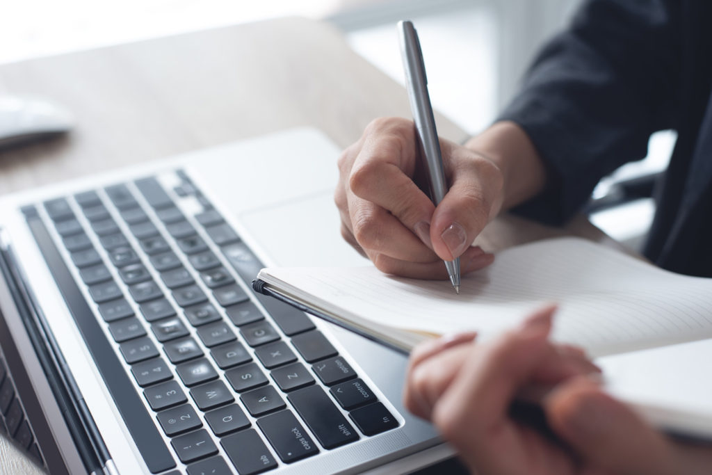 Eine Person schreibt mit einem Stift in einem Notizbuch neben einem Laptop, was ein Symbolbild für Reflexion, Journaling und kritisches Denken ist.