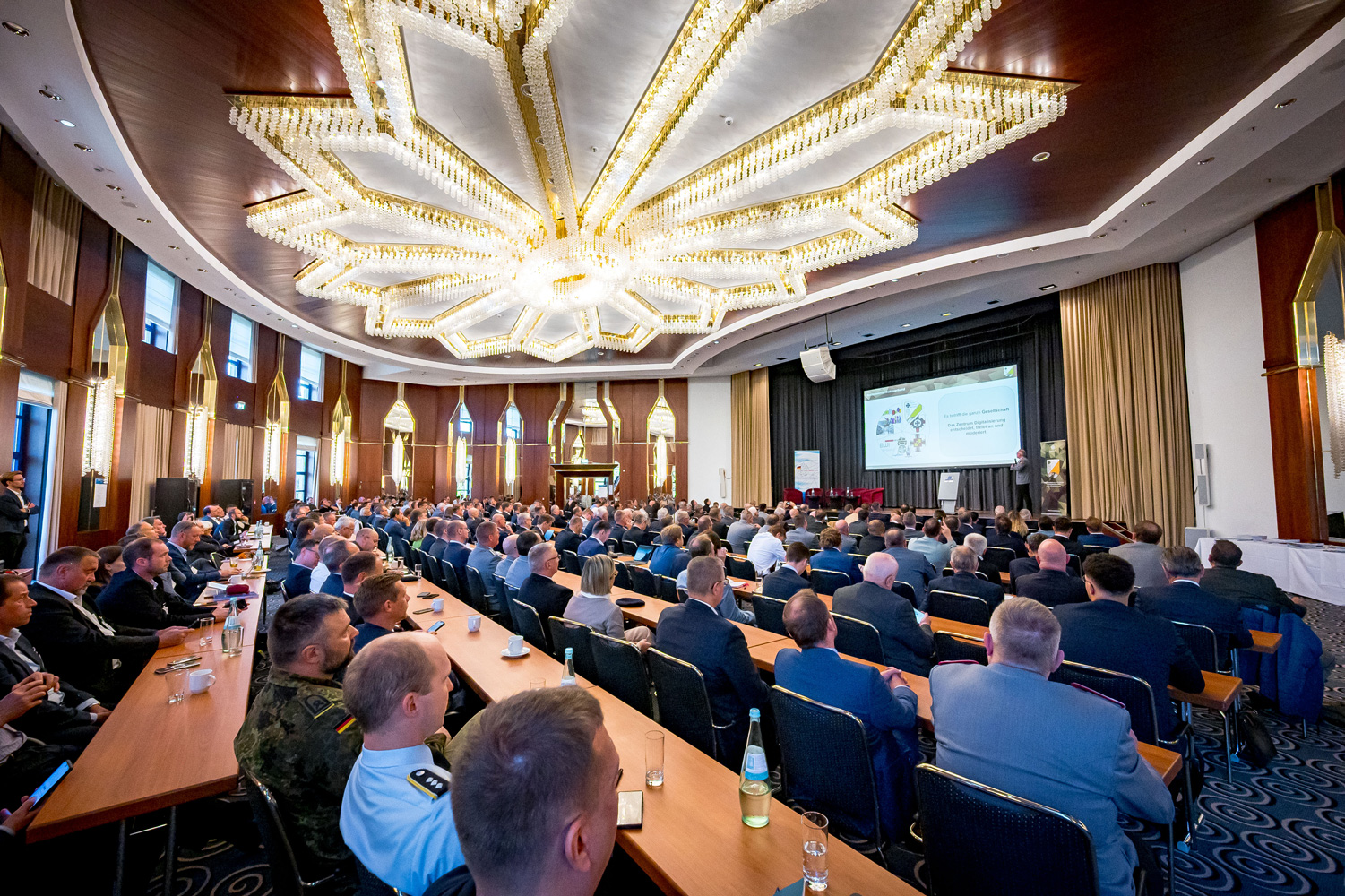 Hier sehen Sie eine Innenaufnahme des Konferenzraums des ersten Bonner IT-Dialogs. Zu sehen sind sowohl das Publikum als auch die Präsentation von Brigadegeneral Bernd Stingl vom Zentrum Digitalisierung der Bundeswehr zur Fähigkeitsentwicklung im Cyber- und Informationsraum.